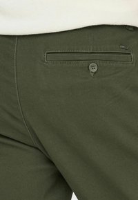 Pantalones verde oliva con una textura suave, que presentan un bolsillo trasero de diseño horizontal y un detalle de botón visible.