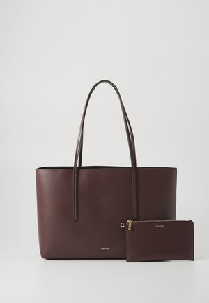 Sac cabas Calvin Klein marron foncé avec deux poignées et une pochette zippée assortie, le tout sur un fond uni clair.