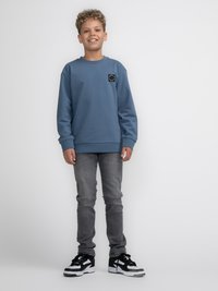 Sudadera azul con cuello redondo y un parche de logo negro, combinada con jeans gris ajustados y zapatillas negras y blancas.