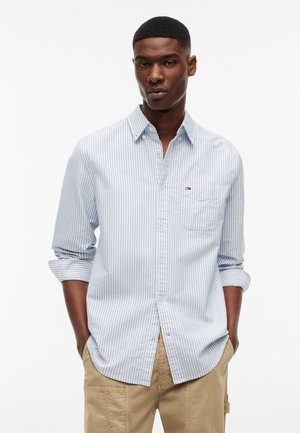 Νεαρός μαύρος άνδρας που φοράει πουκάμισο με κάθετες ρίγες σε ανοιχτό μπλε και λευκό, με κουμπιά και μανίκια γυρισμένα, καθώς και μπεζ παντελόνι, μπροστά σε απλό λευκό φόντο.