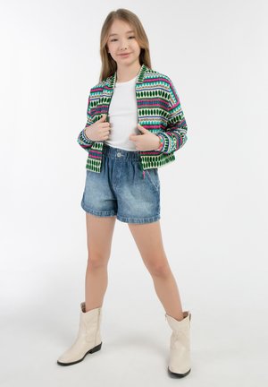 Giacca bomber a maglia multicolore con un motivo geometrico, abbinata a pantaloni shorts in denim chiaro e stivali da cowboy bianchi. Texture liscia, stile casual.