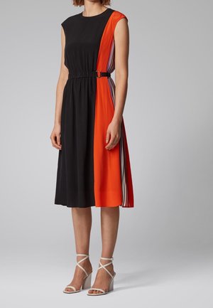 Vrouw die een mouwloze midi-jurk draagt met een zwarte voorkant, feloranje zijpaneel, gestreepte accenten en witte sandalen met hoge hakken.