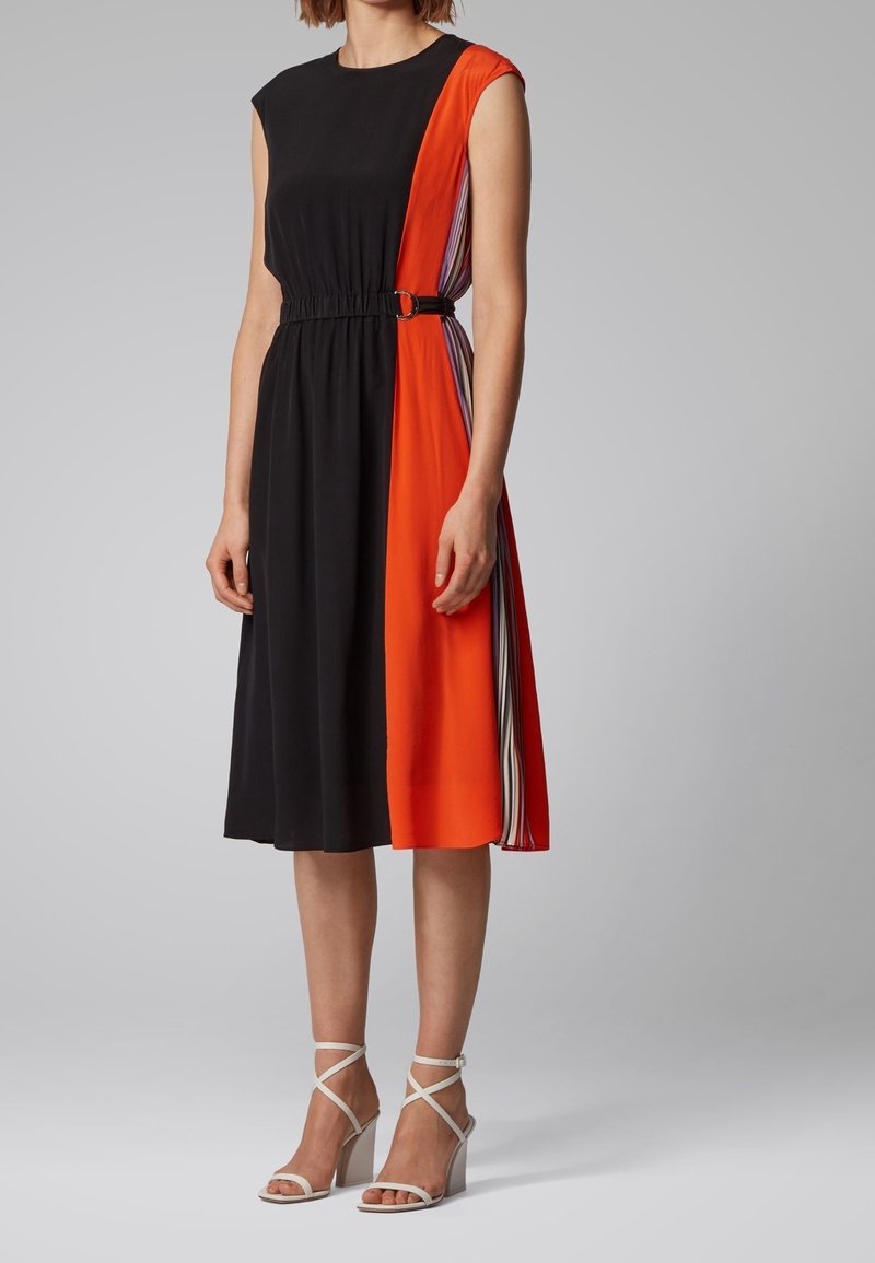 Femme portant une robe midi sans manches avec un devant noir, un panneau latéral orange vif, des accents rayés et des sandales blanches à talons hauts.