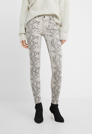 Vrouw die beige en grijs slangenprint strakke broek draagt, lichtbeige trui en zwarte enkelhoge laarzen met hak, staand op een witte achtergrond.