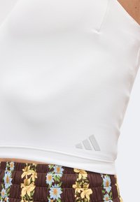 Top de sport blanc court avec un petit logo Adidas près de l’ourlet, porté avec un pantalon marron à motif floral.