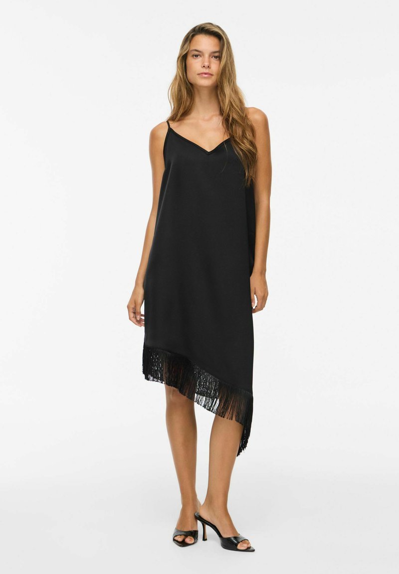 Femme portant une robe noire sans manches avec un ourlet à franges et des sandales à talons ouvertes noires, debout devant un fond blanc.