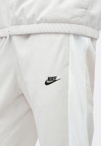 Pantalones de chándal Nike de color gris claro con un logo negro en el lateral. La tela es suave, con una textura ligeramente brillante y un diseño entallado.