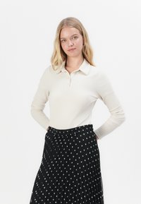 Polo shirt a maniche lunghe a coste di colore crema, abbinato a una gonna plissettata nera con pois bianchi. Colletto semplice e vita elasticizzata.