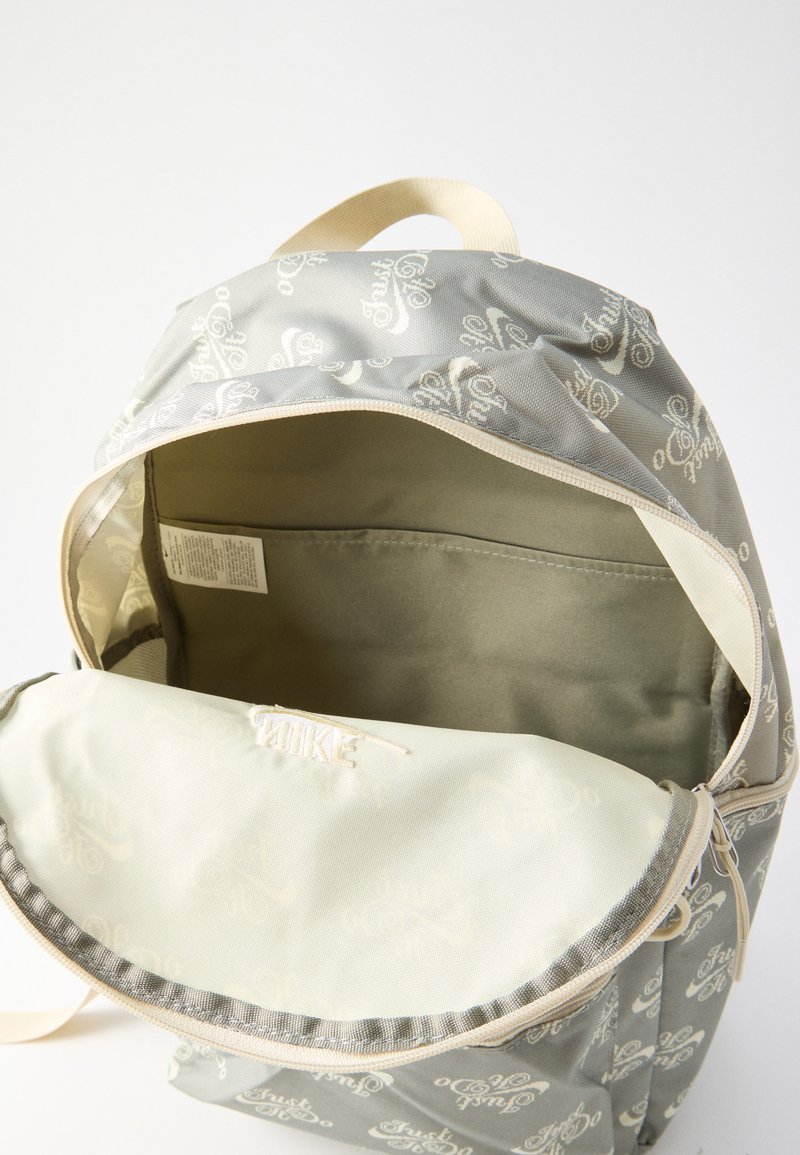 Grauer Rucksack mit weißem sich wiederholenden Schriftmuster, geöffnet, um das leere Hauptfach und das innere Etikett auf beigem Hintergrund zu zeigen.
