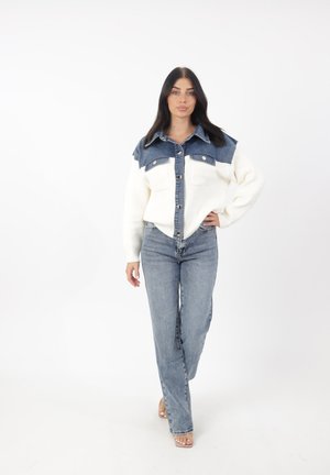 Chemise à manches longues blanche en denim, avec un col bleu et deux poches avant. Portée avec un jean droit et des sandales.