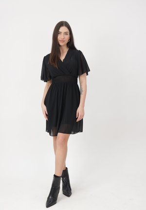 Mujer joven con cabello largo y oscuro, vestida con un vestido negro hasta la rodilla con mangas abullonadas y botas negras hasta el tobillo, de pie sobre un fondo blanco.