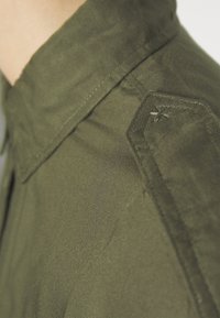 Nahaufnahme einer olivgrünen Stoffschulter mit einer aufgenähten Epaulette, die eine kleine Kreuzstich-Detailierung aufweist.