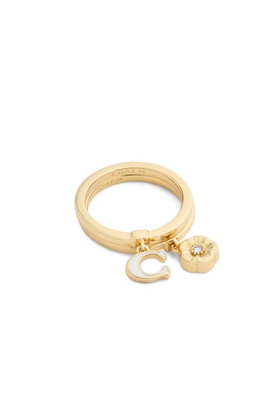 Bague dorée avec deux breloques : un "C" en émail blanc et une fleur avec un détail central en cristal, sur fond blanc.