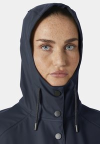 Vrouw met blauwe ogen en sproeten draagt een marineblauw regenjas-capuchon die is vastgemaakt met drie zwarte knopen en grijze trekkoorden.