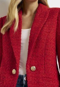 Femme portant un blazer rouge texturé à boutons dorés sur un haut blanc côtelé et un jean bleu, montrant le haut du torse et le cou.