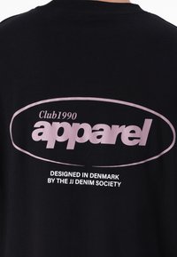 T-shirt en coton noir avec un grand logo ovale rose portant le texte "Club1990 apparel" et un plus petit texte blanc en dessous, indiquant le design et la marque.