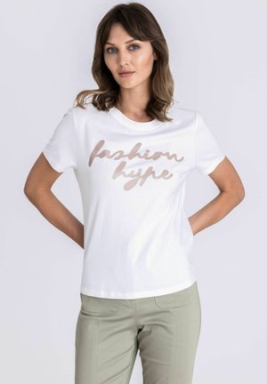 Vrouw met schouderlang bruin haar, draagt een wit T-shirt met de tekst "fashion hype" en lichtgroene broek, handen rustend achter haar rug.