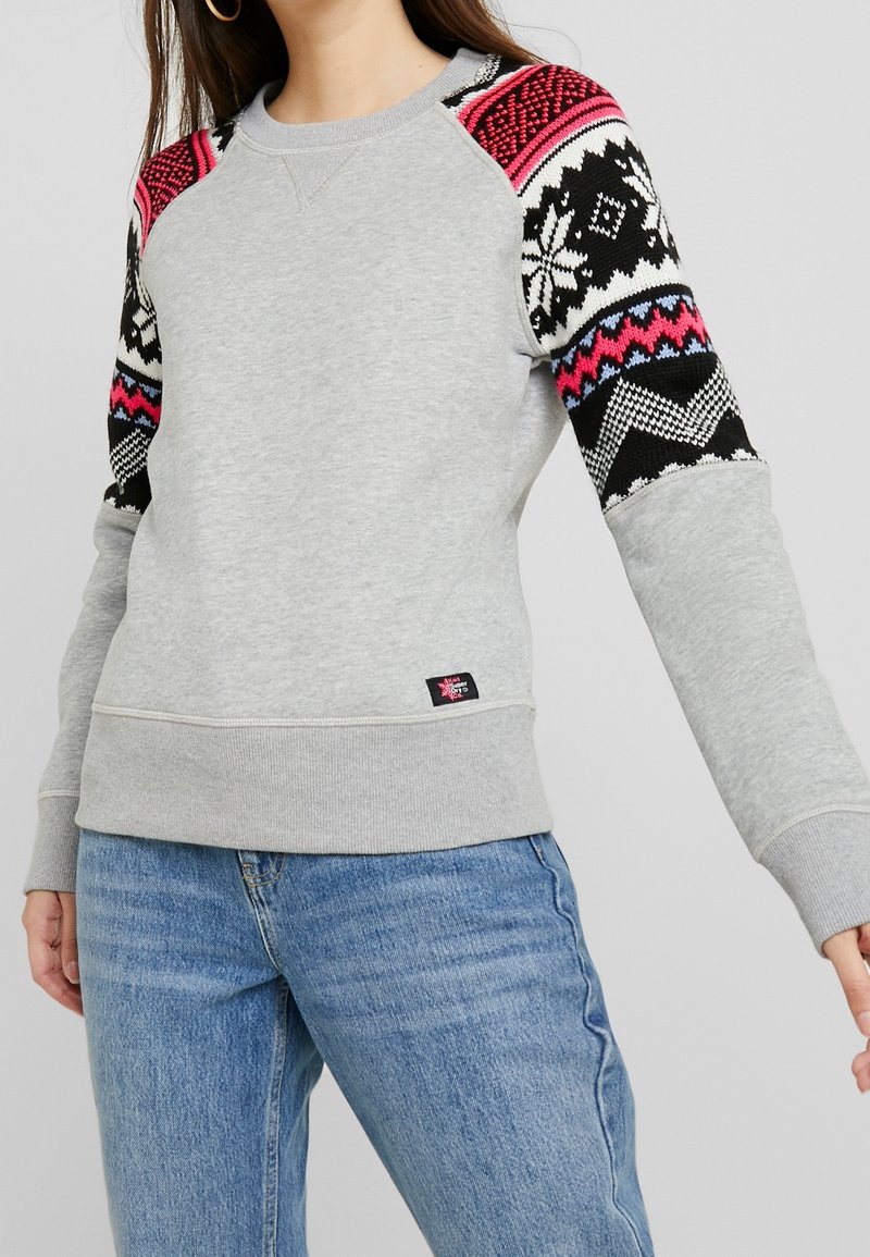 Femme portant un sweat-shirt gris avec des manches en tricot à motifs noirs, blancs et roses et un jean bleu sur un fond uni.