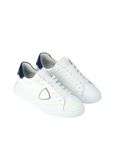 Witte lage sneakers met marineblauwe suède hieltabs, witte veters en een gestikt patchdetail aan de zijkant, weergegeven tegen een witte achtergrond.