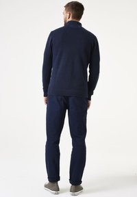 Dunkelblauer Strickcardigan mit hochgeschlossenem Kragen, strukturiertem Muster und geripptem Saum. Kombiniert mit dunklen Hosen und grauen Turnschuhen.