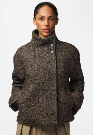 Une femme présente un manteau en tweed marron foncé, à col montant et gros boutons, sur un fond blanc uni.