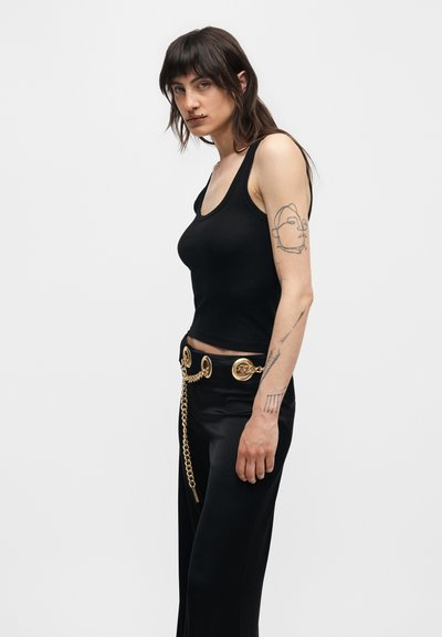 Γυναίκα με μαύρο αμάνικο τοπ και παντελόνια με χρυσής αλυσίδας ζώνη, που δείχνει αφηρημένη τατουάζ προσώπου στον αριστερό πάνω βραχίονα, στέκεται μπροστά σε λευκό φόντο.