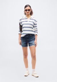 Γυναίκα που φοράει ριγωτό μακρυμάνικο πουκάμισο, τζιν σορτς, λευκές κάλτσες, αθλητικά παπούτσια, γυαλιά ηλίου και κρατά μια μικρή τσάντα ώμου με λουρί αλυσίδα.