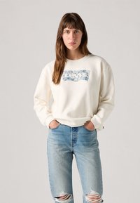 Sweatshirt krem boje s cvjetnim grafičkim logotipom Levi's, okrugli izrez, duge rukave i opušteni kroj, u kombinaciji s svijetloplavim ispucanim trapericama.