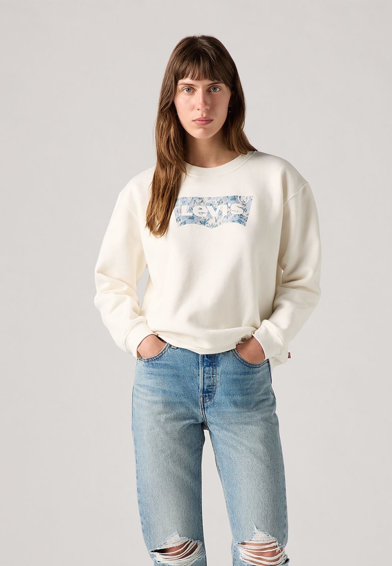 Sweatshirt krem boje s cvjetnim grafičkim logotipom Levi's, okrugli izrez, duge rukave i opušteni kroj, u kombinaciji s svijetloplavim ispucanim trapericama.