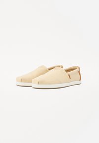 Des chaussures slip-on beiges en tissu texturé, avec une semelle en caoutchouc blanche et des accents en cuir marron sur le talon et sur le côté.