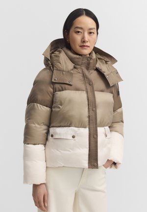 Frau trägt eine color-blocked Steppjacke in Beige, Braun und Creme mit hohem Kragen und Druckknopfdetails, steht mit einer Hand in der Tasche.