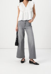 Vrouw in een witte blouse met korte mouwen, hoge taille wijdvallende grijze jeans, zwarte studschoenen en een zwarte handtas.