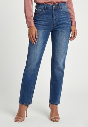Vrouw draagt blauwe jeans met middenhoge taille en nude hoge hak sandalen, staand tegen een effen achtergrond.