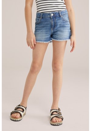 Dítě oblečené v modrých džínových kraťasech, pruhovaném tílku a černo-bílých sandálech, stojící na béžovém pozadí.