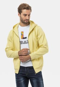Geel ritsvest met een zachte textuur, voorzien van een zak aan de voorkant en een capuchon met koord, gedragen over een witte shirt met kleurrijke grafische print.