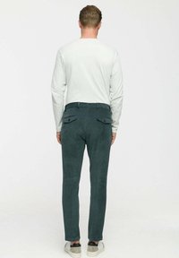 Sötétzöld kordbársony nadrágot, fehér hosszúujjú inget és szürke tornacipőt viselő férfi, háttal áll egy egyszínű háttér előtt.