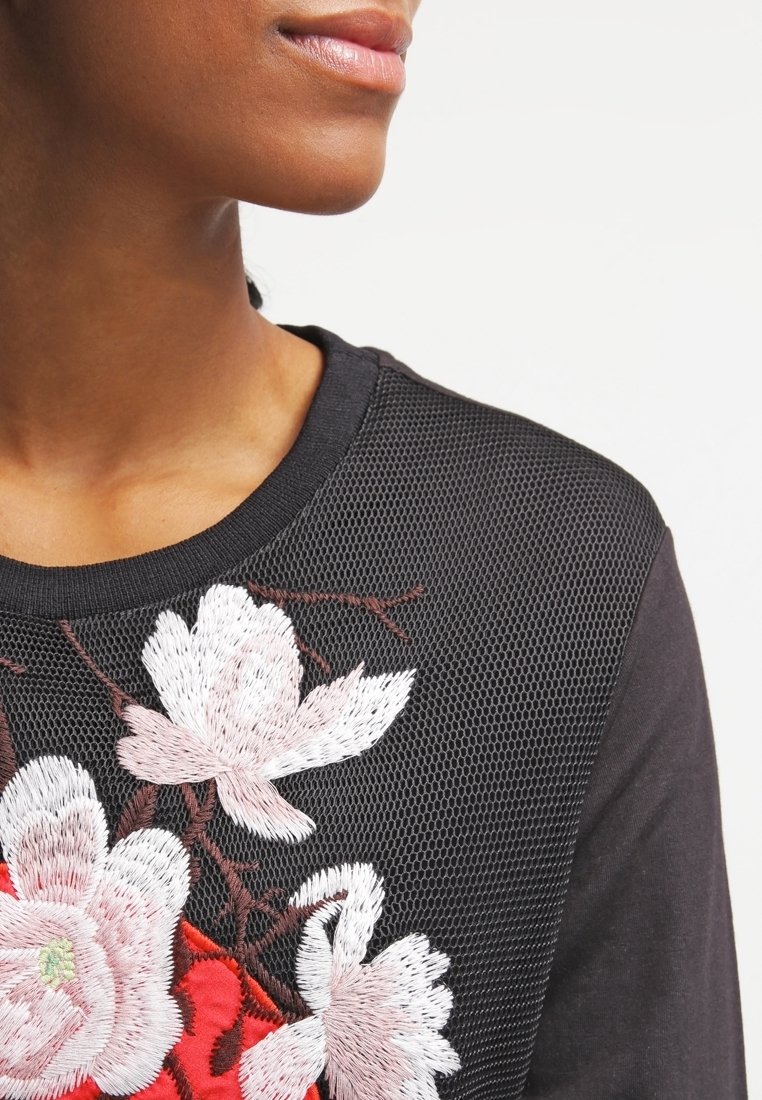 Camisa negra con una sección superior de rejilla que presenta bordado floral en rosa y rojo, con acentos verdes y un cuello redondo.