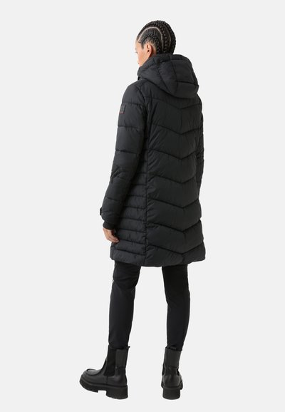 Schwarze Pufferjacke mit Kapuze, gestepptem Design, langen Ärmeln und Seitentaschen. Getragen über schwarzen Hosen und klobigen schwarzen Stiefeln.