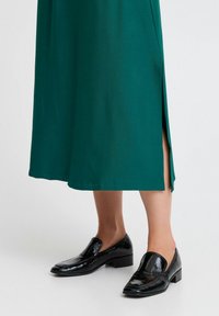 Robe longue verte avec une fente sur le côté, en tissu lisse, associée à des mocassins noirs brillants avec un petit talon carré.