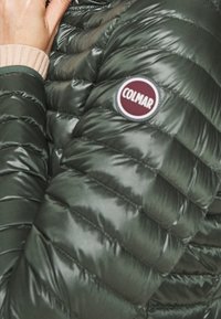 Zelená péřová bunda s prošívanou texturou, lesklou tkaninou a kulatou nášivkou Colmar na rukávu. Viditelné detaily ribbovaného úpletu na manžetách.