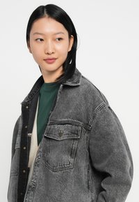 Jovem mulher com cabelo preto liso, vestindo uma jaqueta de denim preta desbotada sobre uma blusa verde, olhando levemente para a sua esquerda em um fundo branco.