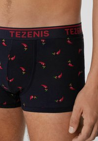 Černé boxerky s těsným střihem, zdobené vzorem červených papriček. Pas je červený a nese značku "TEZENIS" tučnými písmeny.