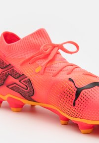 Scarpino da calcio rosa brillante con tomaia in rete, superficie texturizzata, dettagli neri e suola arancione a contrasto. Presenta un design tradizionale con lacci.