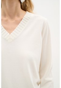 Weißer Pullover mit V-Ausschnitt, ausgestattet mit einem gerippten Kragen und einer weichen Textur. Das Design umfasst lange Ärmel mit einer lässigen Passform.