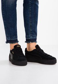 Le sneaker platform nere in suede presentano un upper testurizzato, lacci piatti, il logo PUMA e una suola in gomma rigata. Jeans con orli sfrangiati visibili.