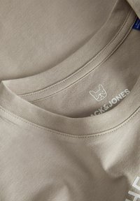 Svijetlosmeđi pamučni T-shirt, okrugli izrez, vidljivi šavovi i bijeli tiskani logo sa siluetom psa na unutarnjoj etiketi.