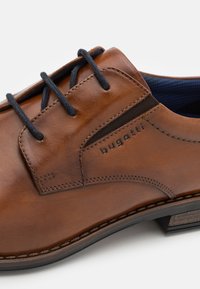 Brown leather shoe with a smooth finish, features blue laces, embossed logo, and contrasting textured panel on the side.