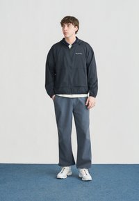 Νεαρός άνδρας που στέκεται σε μπλε χαλί, φορώντας μαύρο μπουφάν Columbia, μπλε παντελόνι, λευκά αθλητικά παπούτσια με πολύχρωμες λεπτομέρειες, με τα χέρια στις τσέπες.