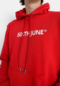 Röd bomullshuvtröja med en ficka fram, snörning med metallspetsar och "SIXTH JUNE"-logotyp i vit brodering på bröstet.