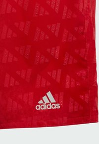 Crvena sportska dres s teksturiranim kapljicama vode. Ima istaknuti bijeli Adidas logo i geometrijski uzorak preko tkanine.
